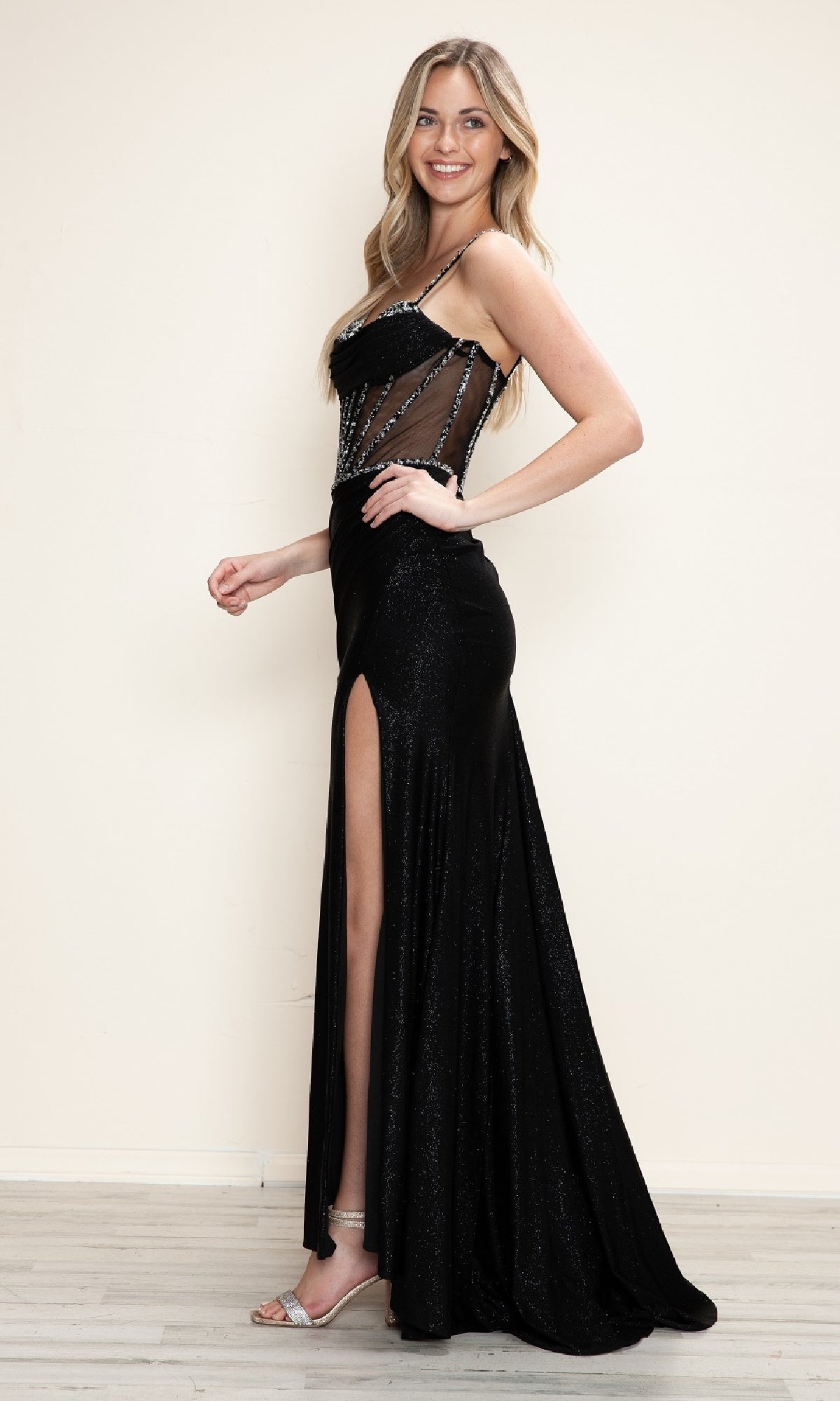 A woman poses sideways against a light background, wearing the Sheer Beaded-Corset Long Prom Dress: Poly USA 9750—featuring a black beaded sheer bodice, thigh-high slit, and paired with silver high-heeled sandals.